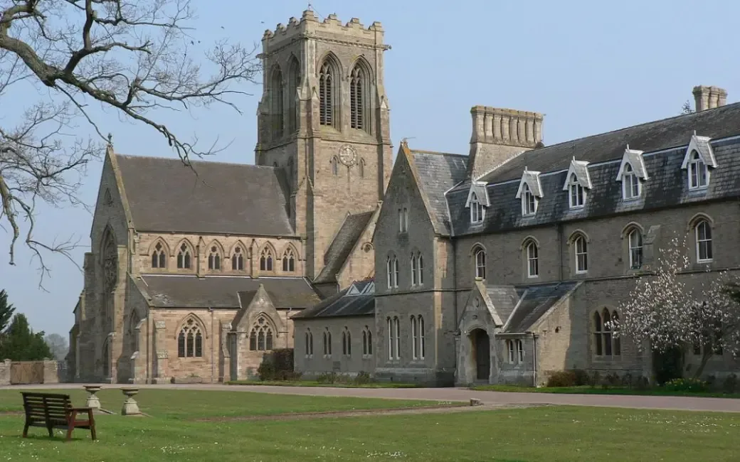 Large stone building with tall arched windows and a central clock tower, resembling a historic church or abbey, surrounded by green lawns and a tree branch in the foreground.